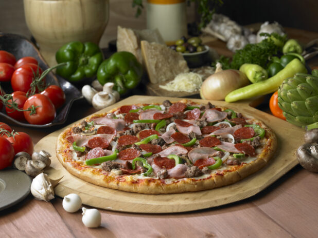 A Whole Gourmet Deluxe Pizza with Sliced Ham, Ground Beef, Pepperoni and Sliced Vegetables on a Wooden Pizza Peel on a Wooden Table