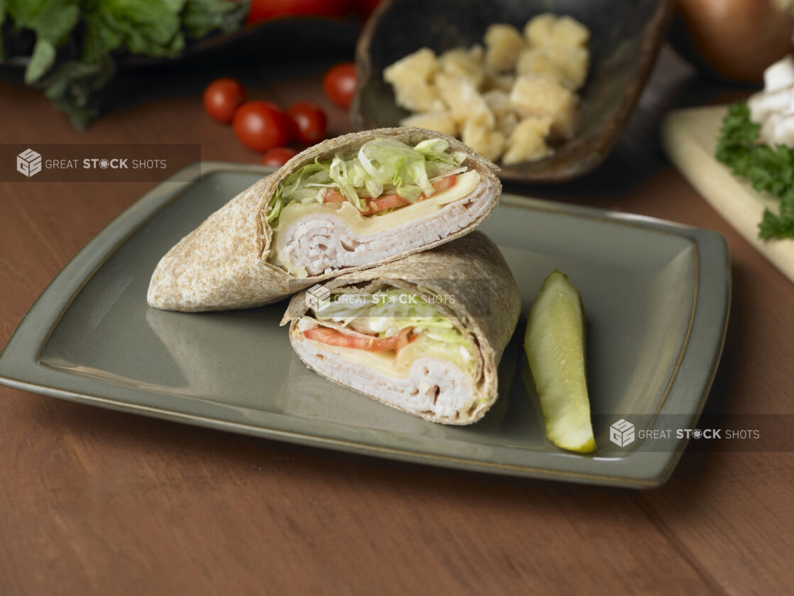 Turkey and Swiss in a Whole Wheat Wrap with Fresh Vegetables and a Pickle Spear on a Grey Ceramic Dish on a Wooden Table