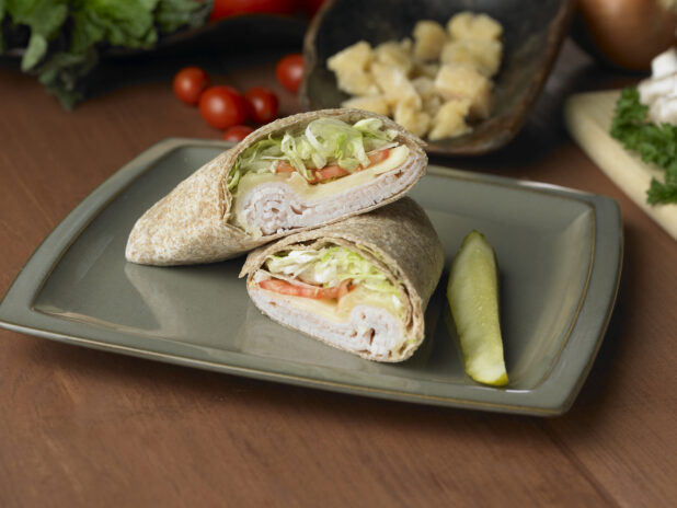 Turkey and Swiss in a Whole Wheat Wrap with Fresh Vegetables and a Pickle Spear on a Grey Ceramic Dish on a Wooden Table