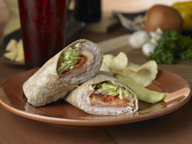 Turkey, Ham and Swiss in a Whole Wheat Wrap with Fresh Vegetables and a Pickle Spear on a Brown Ceramic Dish on a Wooden Table