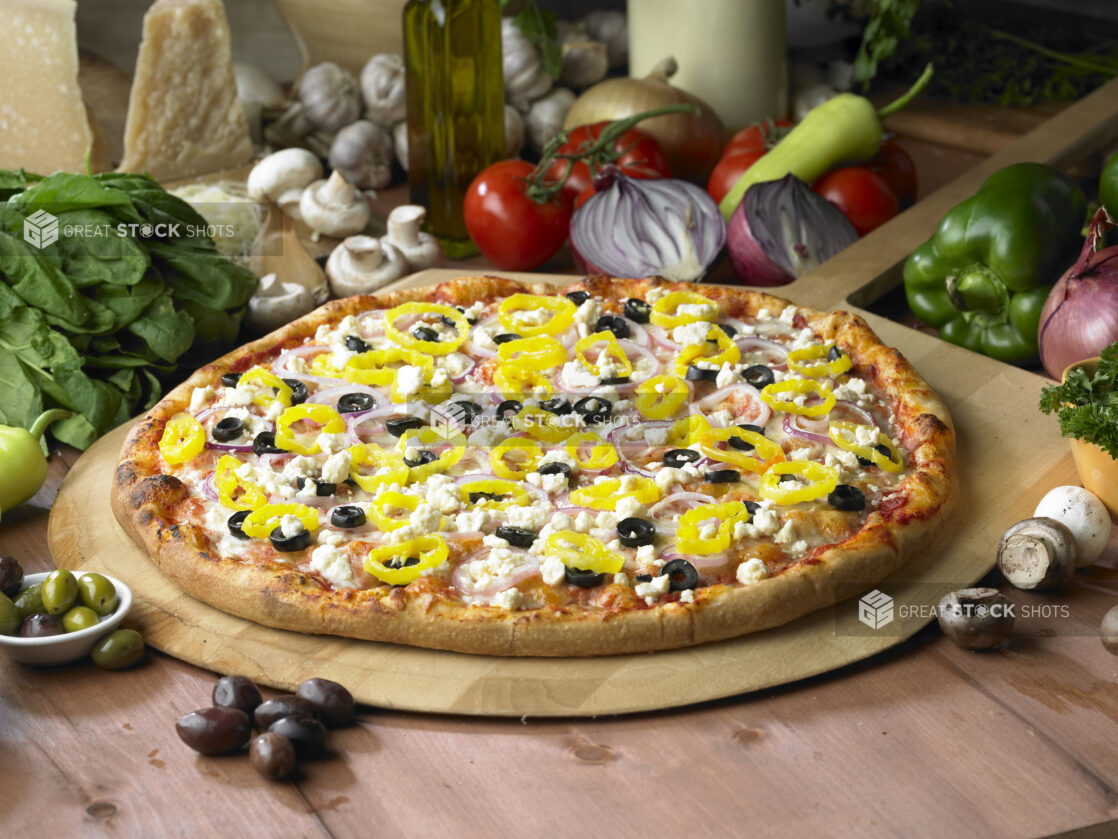 Whole Greek Pizza with Sliced Black Olives, Hot Banana Peppers, Feta Cheese and Red Onions on a Wooden Pizza Peel in a Indoor Setting