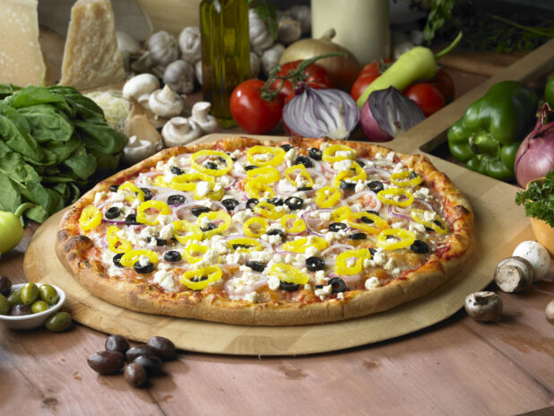 Whole Greek Pizza with Sliced Black Olives, Hot Banana Peppers, Feta Cheese and Red Onions on a Wooden Pizza Peel in a Indoor Setting