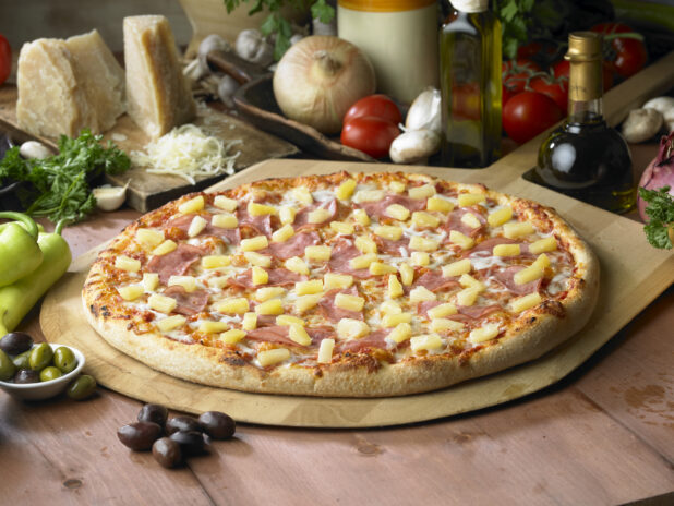 Whole Hawaiian Pizza with Sliced Ham and Pineapple Chunks on a Wooden Pizza Peel in a Indoor Setting