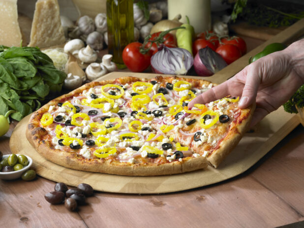 A Hand Grabbing a Slice of Greek Pizza with Sliced Black Olives, Hot Banana Peppers, Feta Cheese and Red Onions on a Wooden Pizza Peel in a Indoor Setting