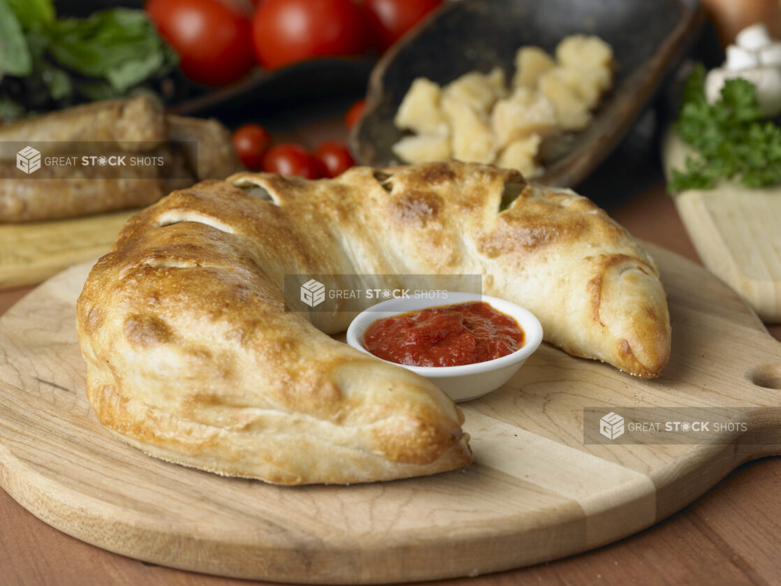 A Whole, Uncut Stromboli with a Side of Marinara Sauce on a Wooden Cutting Board in a Kitchen Setting