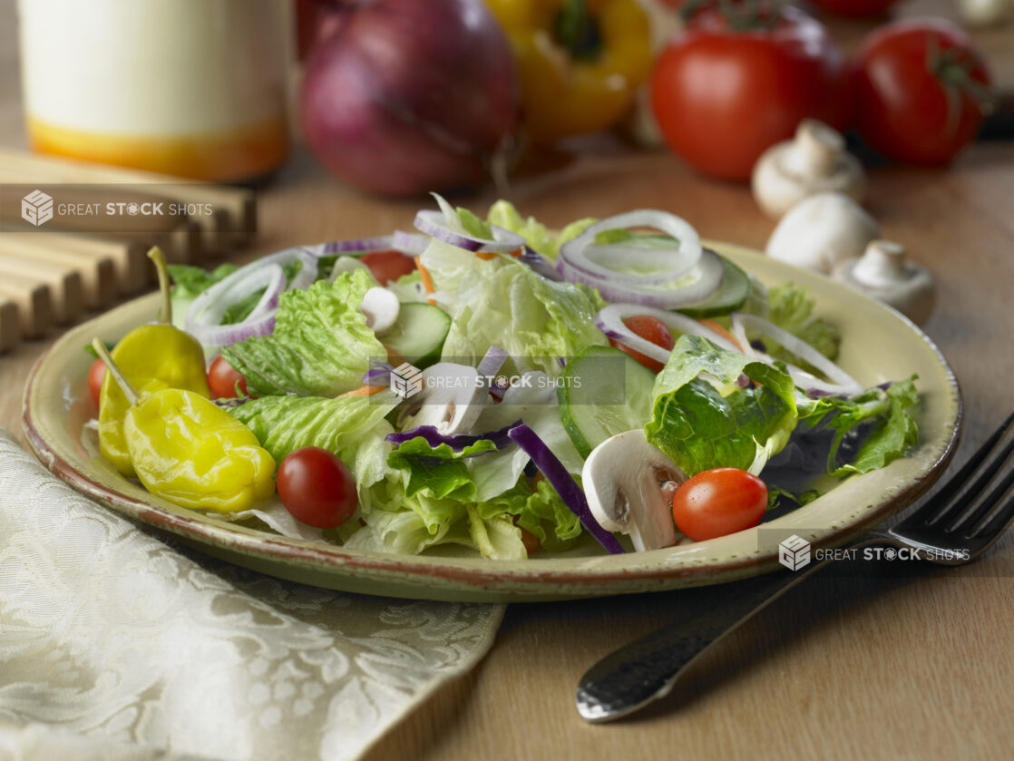 Garden Salad with Sliced Mushrooms, Cucumbers, Red Onions, Cherry Tomatoes and Pepperonicini Peppers in a Beige Ceramic Dish on a Wooden Table