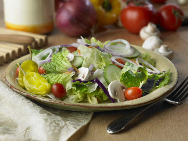 Garden Salad with Sliced Mushrooms, Cucumbers, Red Onions, Cherry Tomatoes and Pepperonicini Peppers in a Beige Ceramic Dish on a Wooden Table