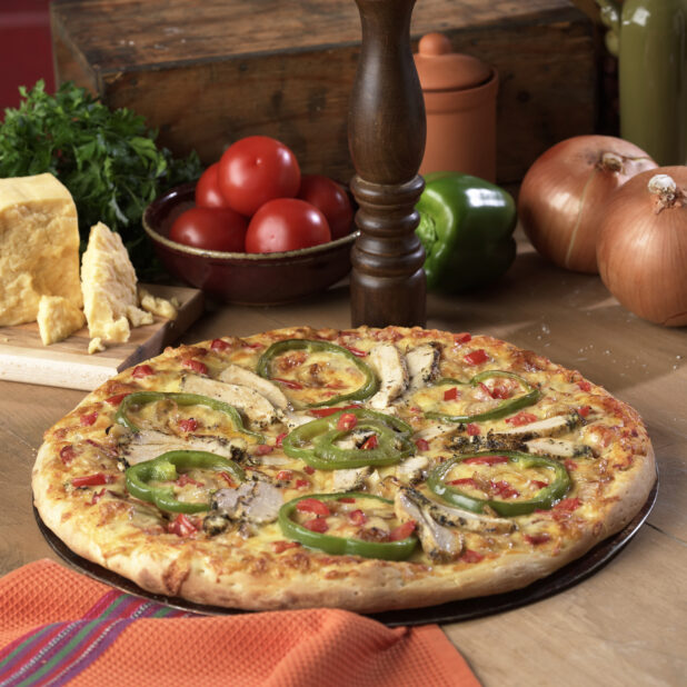 Whole Pizza with Sliced Green Peppers, Diced Tomatoes and Grilled Chicken Toppings on a Wooden Pizza Platter on a Wooden Table in an Indoor Setting