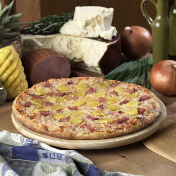 Whole Hawaiian Pizza with Sliced Ham and Pineapple Toppings on a Wooden Pizza Platter on a Wooden Table in an Indoor Setting