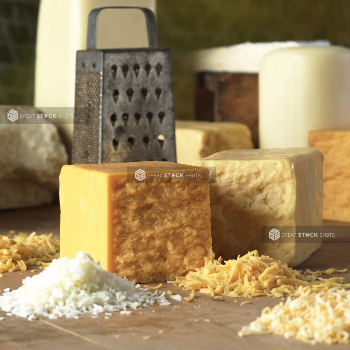 Assorted Blocks of Cheese and Shredded Cheeses on a Wooden Cutting Board with a Metal Cheese Grater in a Kitchen Setting