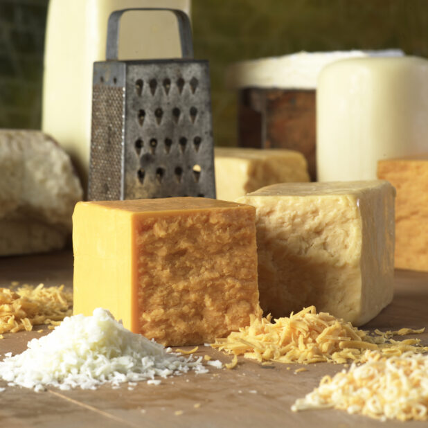 Assorted Blocks of Cheese and Shredded Cheeses on a Wooden Cutting Board with a Metal Cheese Grater in a Kitchen Setting