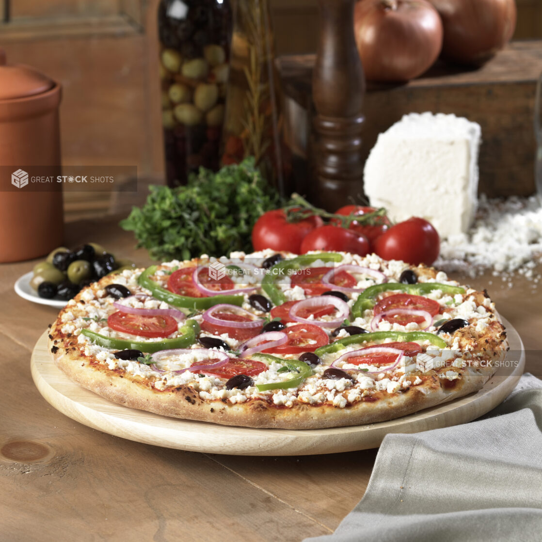 Whole Greek Pizza with Sliced Green Peppers, Tomatoes, Black Olives and Feta Cheese Toppings on a Wooden Pizza Platter on a Wooden Table in an Indoor Setting