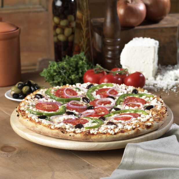 Whole Greek Pizza with Sliced Green Peppers, Tomatoes, Black Olives and Feta Cheese Toppings on a Wooden Pizza Platter on a Wooden Table in an Indoor Setting