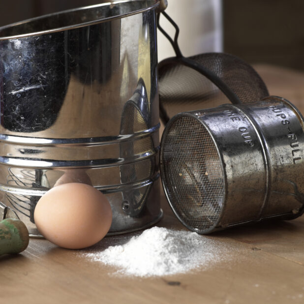 Close Up of Flour Sifters, a Strainer and an Egg on a Wooden Table in a Kitchen Setting