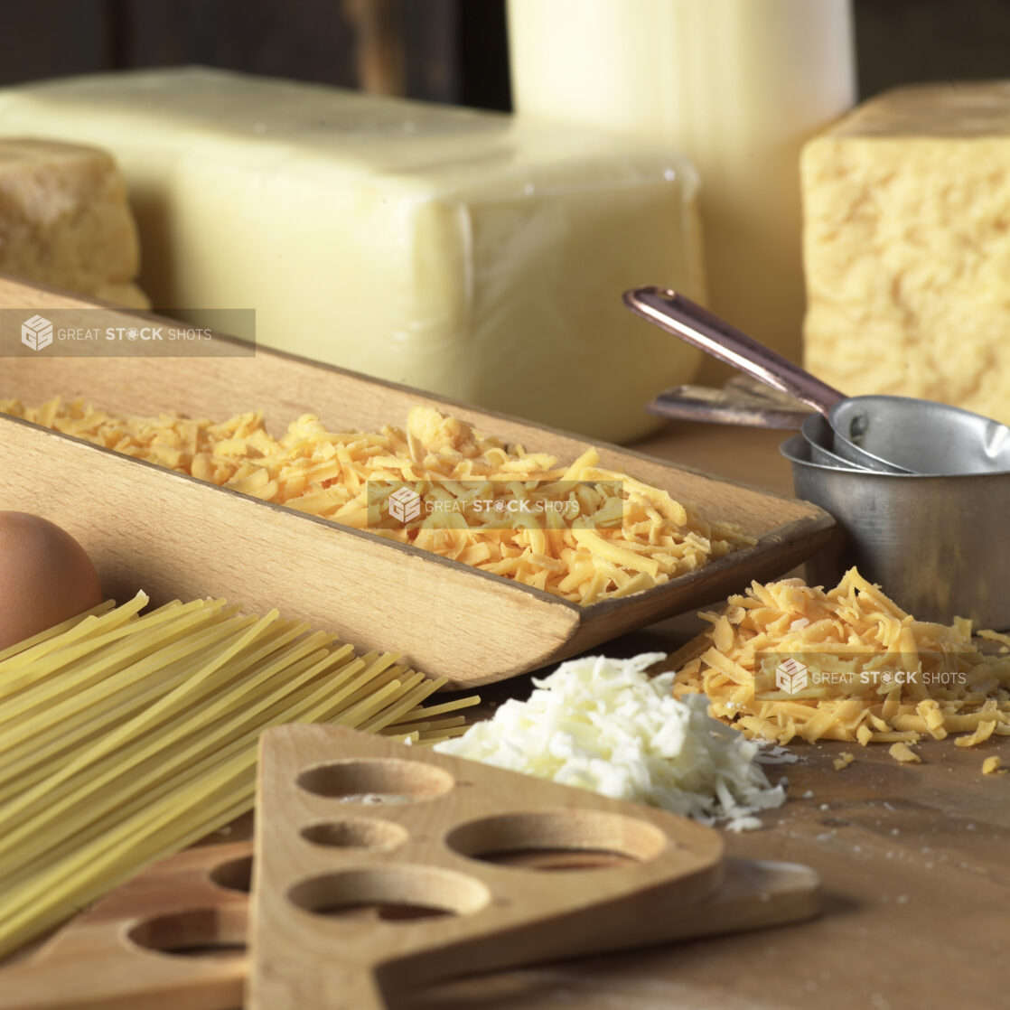 Shredded Cheddar and Mozzarella Cheese, Dry Spaghetti Noodles and Kitchen Utensils on a Wooden Cutting Board in a Kitchen Setting