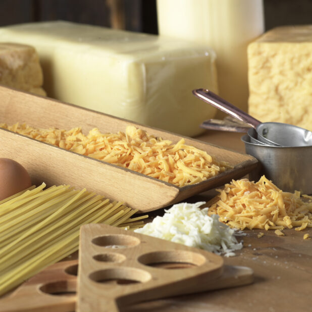 Shredded Cheddar and Mozzarella Cheese, Dry Spaghetti Noodles and Kitchen Utensils on a Wooden Cutting Board in a Kitchen Setting