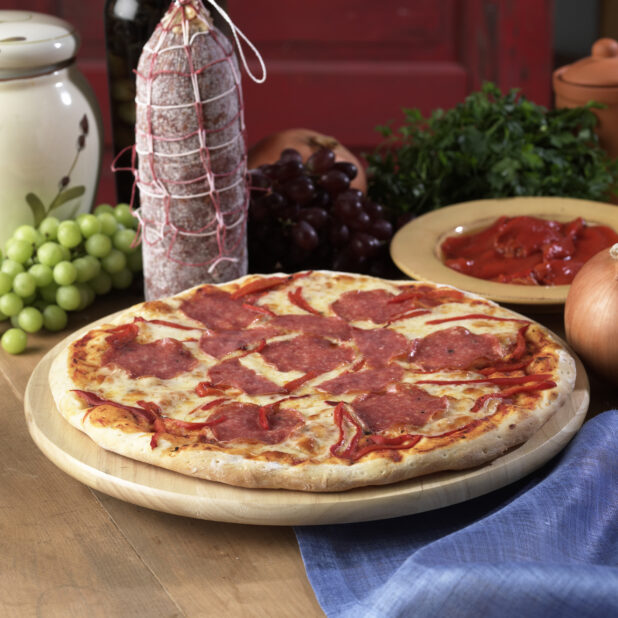 Whole Pizza with Salami and Roasted Red Pepper Toppings on a Wooden Pizza Platter on a Wooden Table in an Indoor Setting
