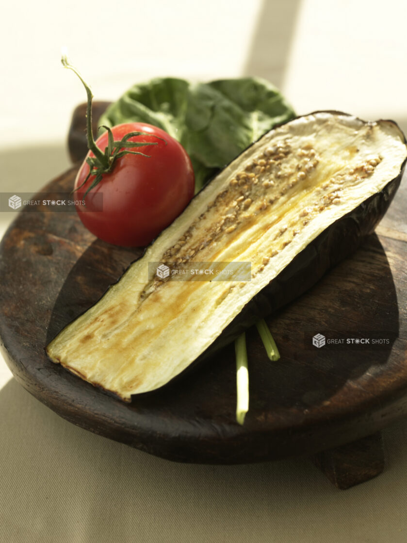 A Whole Vine-Ripened Tomato, Fresh Spinach Leaves and a Roasted Eggplant Half on a Dark Wooden Platter in an Indoor Setting