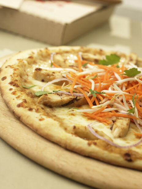 Close Up of a New York-Style Pizza with Asian Sesame Chicken and Vegetable Toppings on a Wooden Pizza Peel in an Indoor Setting