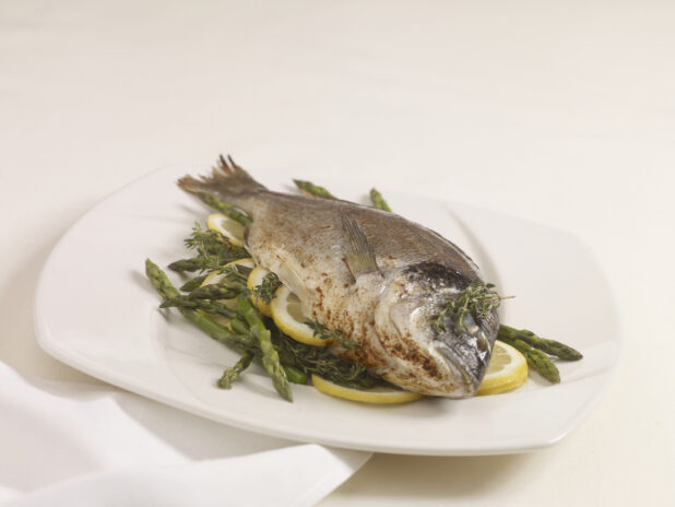 A Whole Roasted Sea Bream with Asparagus, Citrus and Herbs on a White Ceramic Dish