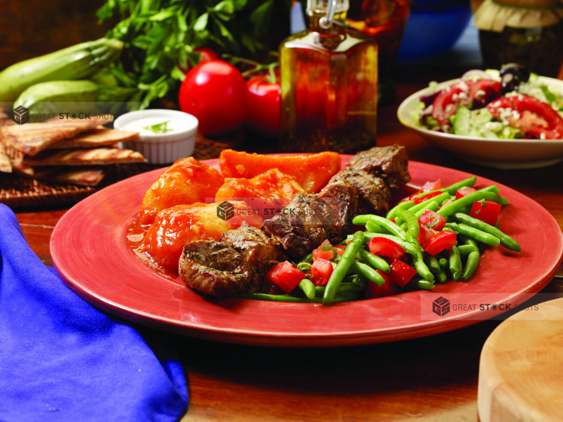 A Combo Plate of Pork Souvlaki, Green Beans and Red Peppers and Roasted Greek Potatoes on a Red Ceramic Dish on a Wooden Table - Variation