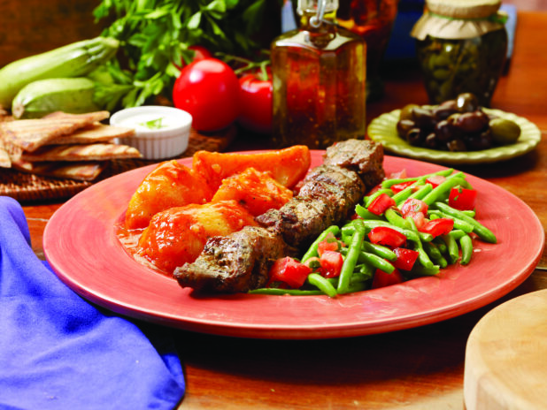 A Combo Plate of Pork Souvlaki, Green Beans and Red Peppers and Roasted Greek Potatoes on a Red Ceramic Dish on a Wooden Table