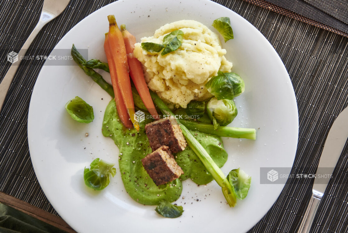 Overhead Shot of Baby Carrots, Mashed Potatoes, Asparagus, Brussels Sprouts and Cubed Steak on a Bed of Green Purée on a White Ceramic Dish