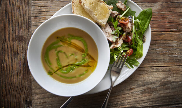 Overhead View of a Butternut Squash and Pea Soup with a Side Salad of Mixed Greens and Grilled Chicken with Sliced Baguettes on a White Ceramic Dish on a Rustic Wooden Table