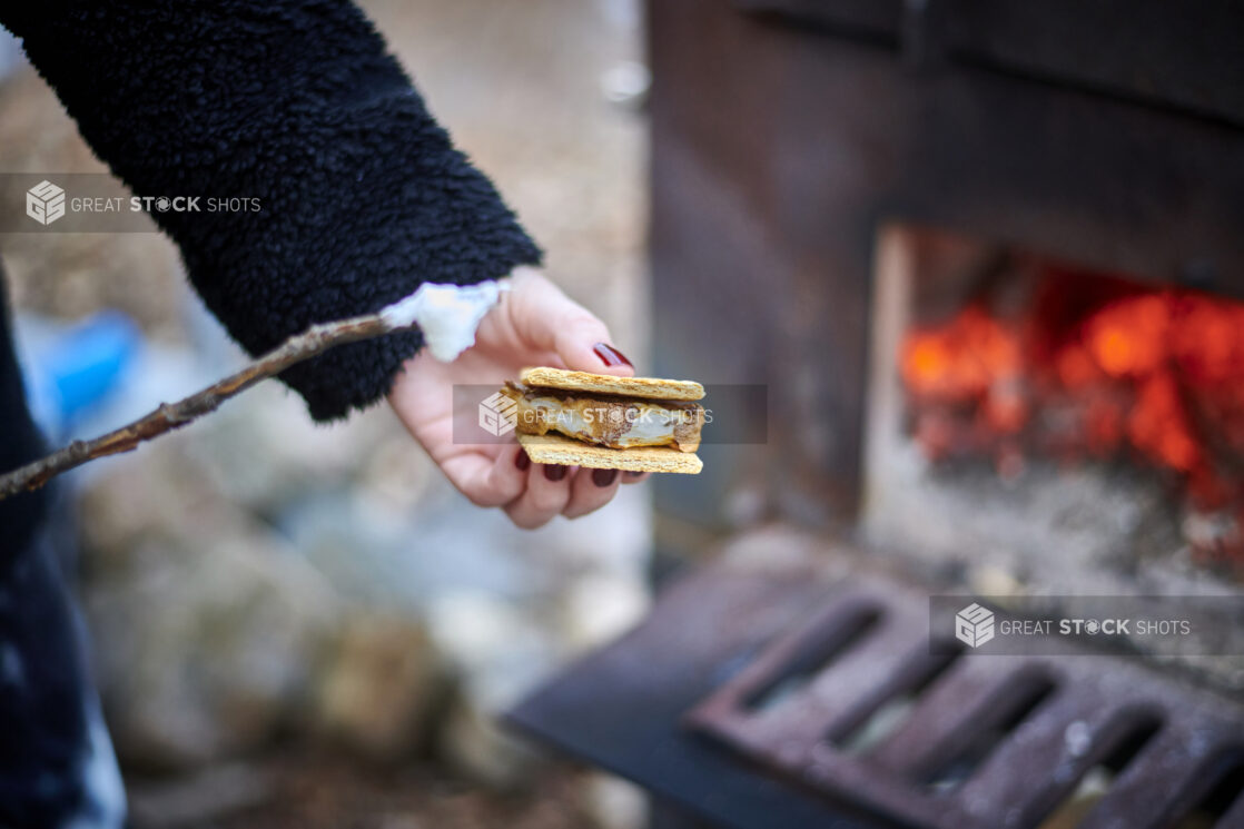 Person Sandwiching a Toasted Marshmallow Between Graham Crackers to Make a S'mores Treat in Front of a Wood-Burning Oven in an Outdoor Setting