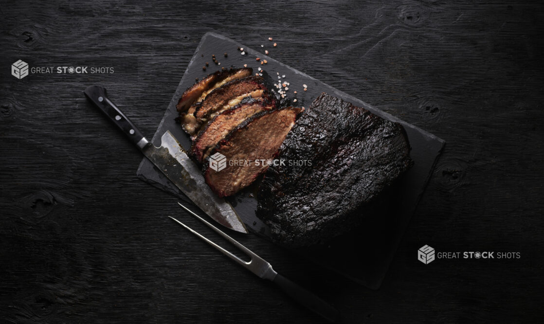 Overhead View of Slices and a Block of Barbecue Beef Brisket on a Slate Platter with Himalayan Pink Salt and Black Peppercorns on a Black Painted Wood Surface