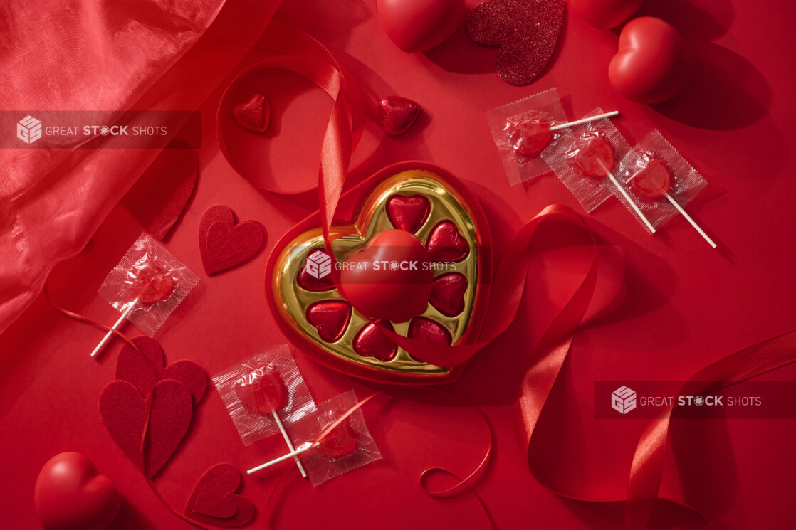 Overhead View of A Box of Heart-Shaped Chocolates Surrounded by Valentine’s Day Decorations on a Red Surface