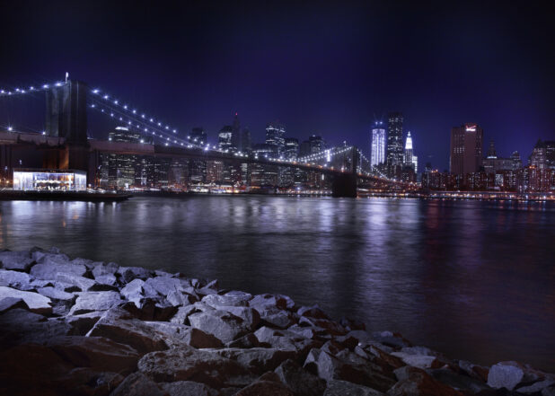 View of the Brooklyn Bridge Lit Up at Night from the Rocky Shores of the East River Seaport in Manhattan, New York City