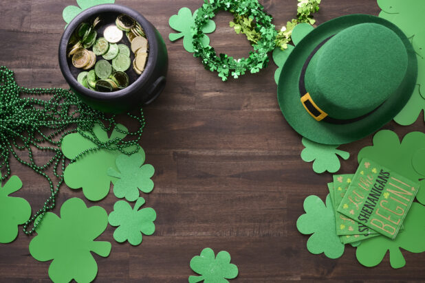 Overhead View of an Assortment of St. Patrick’s Day Decorations – Shamrock Wreaths, Leprechaun Hat, Green Clover Clings and a Pot of Gold on a Dark Wood Background