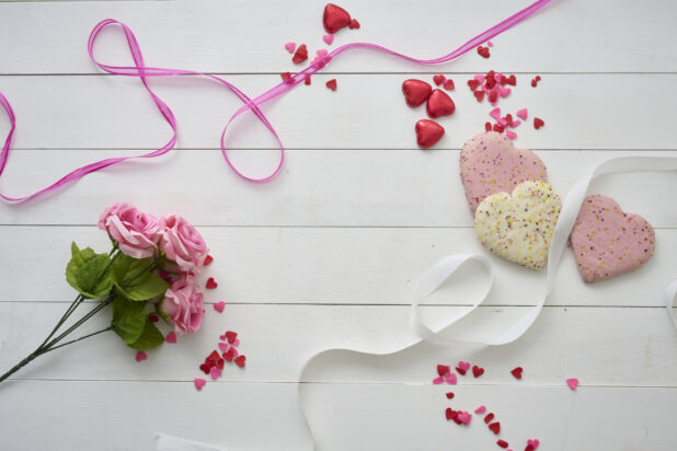 Overhead View of Heart Shaped Cookies, Pink Roses, Satin Ribbons and Heart Confetti on a White Painted Wood Table – Variation 2