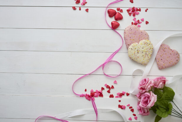 Overhead View of Heart Shaped Cookies, Pink Roses, Satin Ribbons and Heart Confetti on a White Painted Wood Table