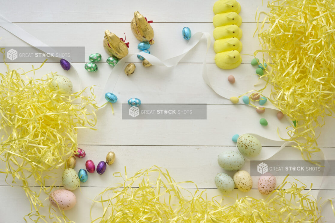 Overhead View of Assorted Chocolate Easter Eggs and Easter-Themed Sweets on a White Painted Wood Table with Yellow Stuffing Paper