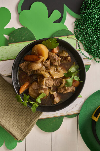 Overhead Shot of a Large Black Bowl of Irish Stew With Carrots, Potatoes and Beef Surrounded by St. Patrick’s Day Decorations