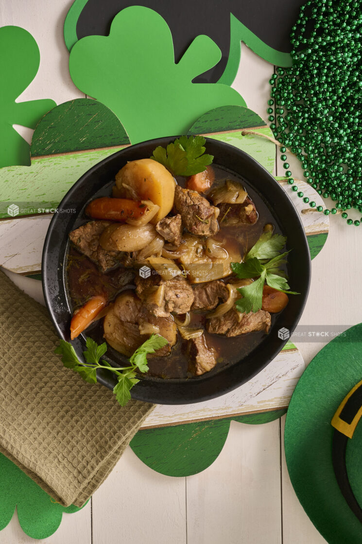 Overhead Shot of a Large Black Bowl of Irish Stew With Carrots, Potatoes and Beef Surrounded by St. Patrick’s Day Decorations