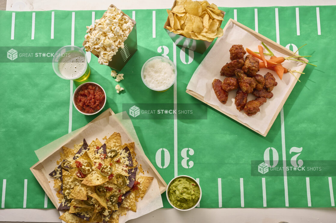 Overhead View of a Spread of Game Time Snacks: Nacho Platter, Chicken Wings, Salsa, Guacamole, Popcorn, Potato Chips and Beer on a Football Field Table Cloth