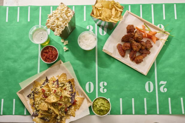 Overhead View of a Spread of Game Time Snacks: Nacho Platter, Chicken Wings, Salsa, Guacamole, Popcorn, Potato Chips and Beer on a Football Field Table Cloth