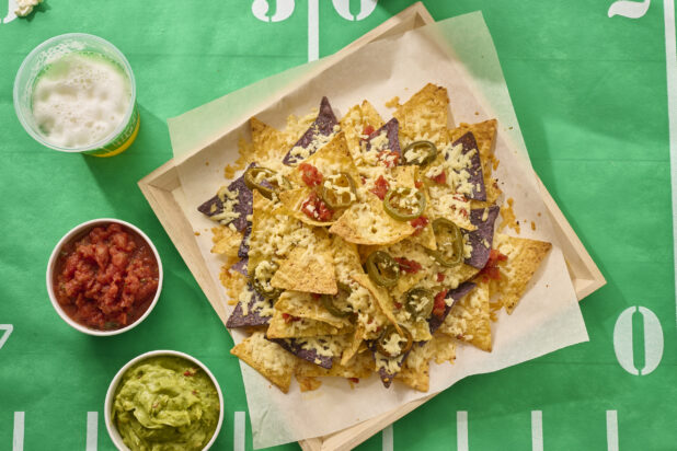 Close Up Overhead View of a Nacho Platter on a Square Wood Tray with Sides of Salsa, Guacamole on a Football Field Table Cloth