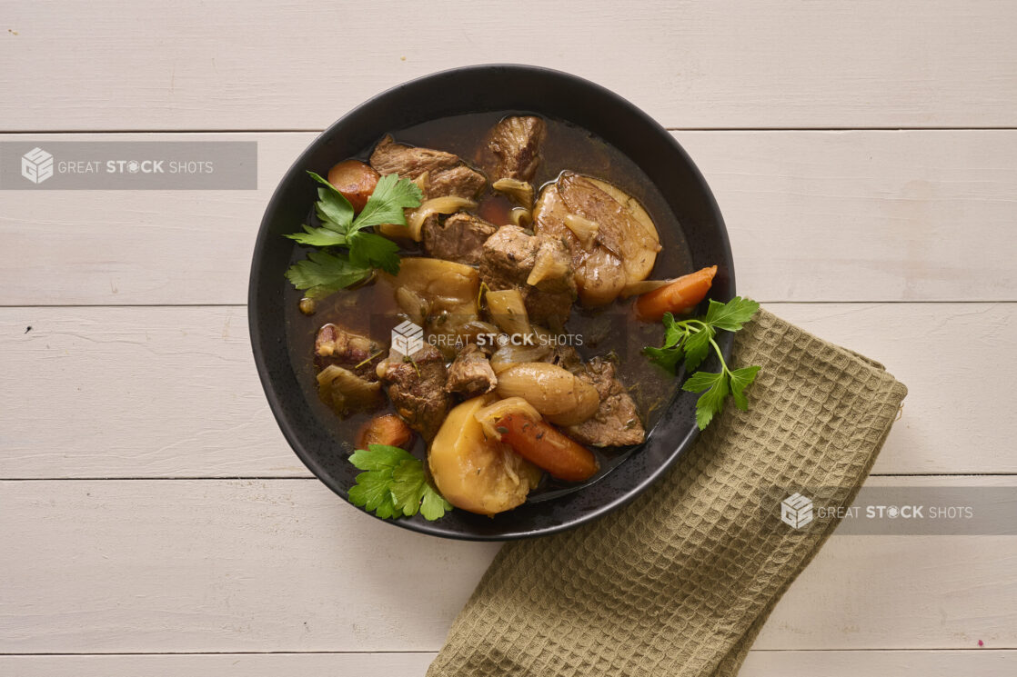 Close Up Overhead View of a Large Black Bowl of Irish Stew With Carrots, Potatoes and Beef on a White Painted Wood Table with a Beige Napkin