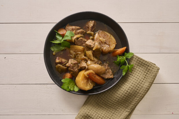 Close Up Overhead View of a Large Black Bowl of Irish Stew With Carrots, Potatoes and Beef on a White Painted Wood Table with a Beige Napkin