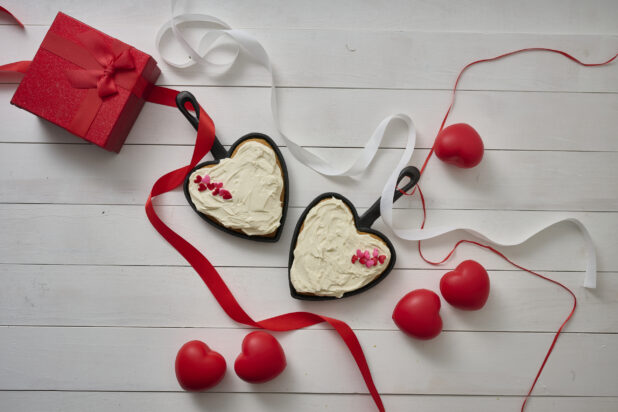 Overhead View of Heart Shaped Cakes with White Frosting and Candy Hearts in Mini Skillet Pans on a White Painted Table with Valentine's Day Decorations