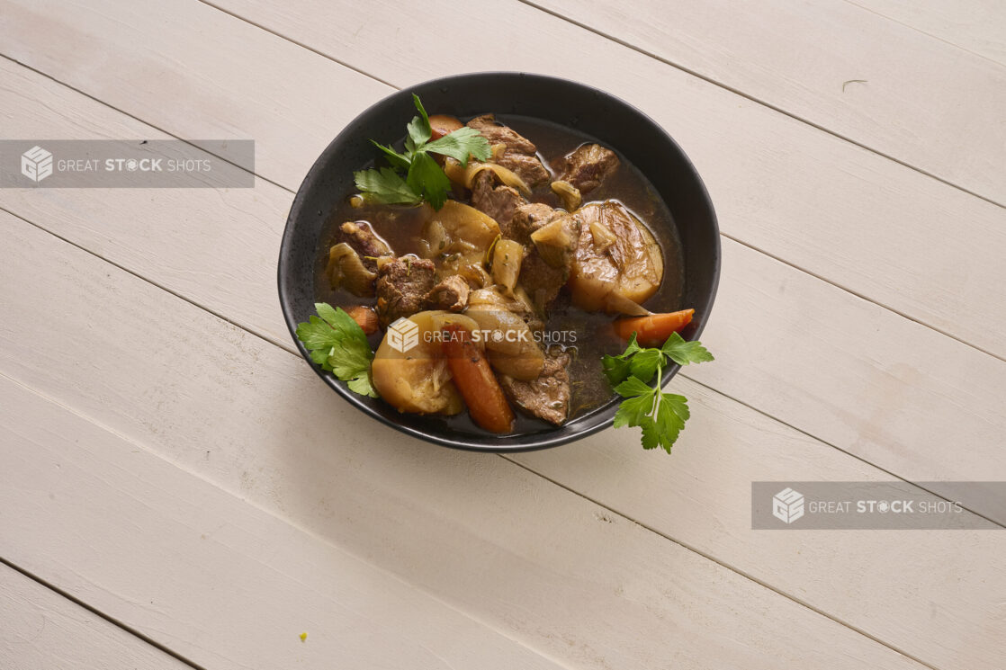 Large Black Bowl of Irish Stew With Carrots, Potatoes and Beef on a White Painted Wood Table