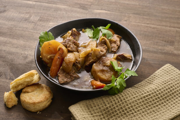Close Up of a Large Black Bowl of Irish Stew With Carrots, Potatoes and Beef on a Dark Wooden Table with Butter Biscuits and a Beige Napkin