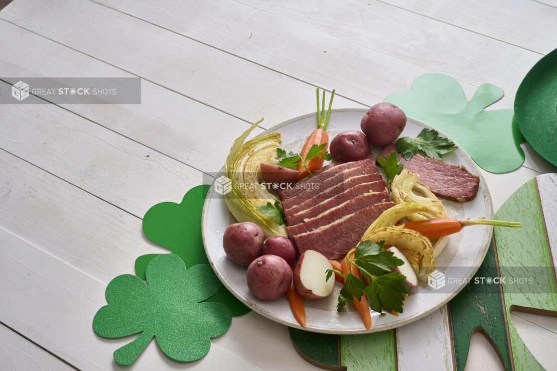 A Plate of Corned Beef and Cabbage with Carrots and Red Skinned Potatoes on a White Painted Wood Table Surrounded by St. Patrick's Day Decorations