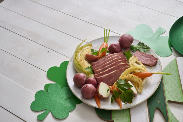 A Plate of Corned Beef and Cabbage with Carrots and Red Skinned Potatoes on a White Painted Wood Table Surrounded by St. Patrick's Day Decorations