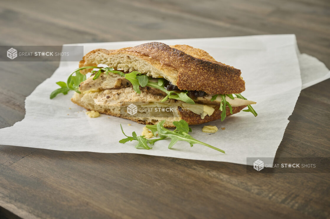 Roasted Chicken Breast and Baby Arugula on Sliced Toasted Baguette on a White Parchment Paper on a Dark Wooden Table