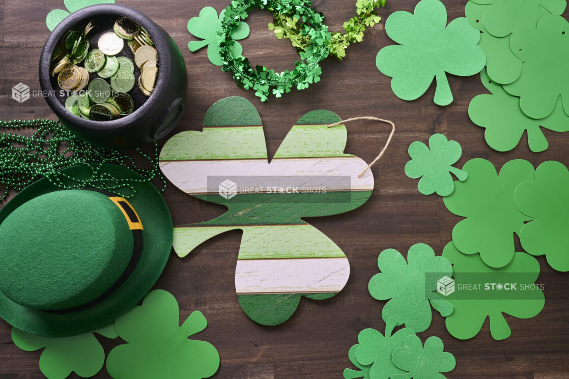 Overhead View of an Assortment of St. Patrick’s Day Decorations – Painted Wood Shamrock, Leprechaun Hat, Green Clover Clings and a Pot of Gold on a Dark Wood Background